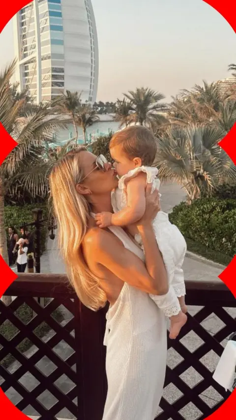A lady holds up and kisses her baby, both dressed in white amid a sunny backdrop