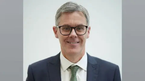 Matthew Warren wearing dark-framed glasses a dark suit, white shirt and green tie looking towards the camera