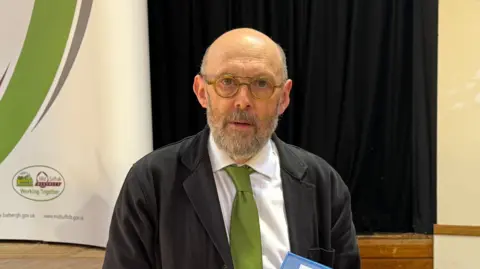 Louise Parker/BBC A head and shoulders image of a man wearing a black jacket over a white shirt and a green tie. He is wearing glasses and looking into the camera. 