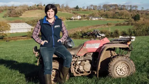 Abi, who has black short hair and wearing a blue gilet above a plaid shirt, sits on a quad bike. Behind her a farm can be seen, with houses in the distance. 