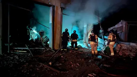 Four people in fire rescue uniforms with torches walk through the rubble of a damaged building in Dnipropetrovsk