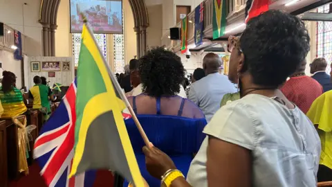 Several people stand inside a church, facing the front. Some are holding the flags of Jamaica and the UK.