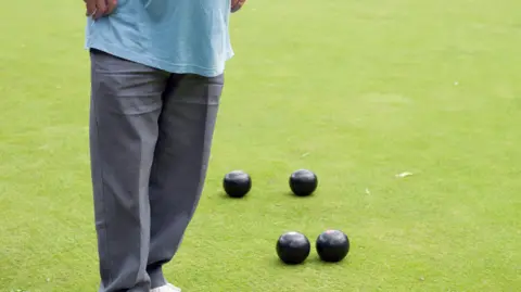 A crown green bowler, pictured from the waist down, stands by four bowls balls on a green. He is wearing a light blue t-shirt and grey trousers.