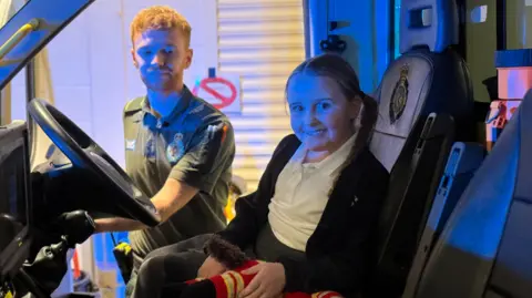 East of England Ambulance Service Bunny-Mae sitting in the driving seat of an ambulance and is being shown how to turn the lights on. She is looking directly at the camera and is smiling.