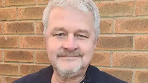 Julie Stevenson David, a man with grey hair and small grey beard, wearing a black T-shirt, looking into the camera.