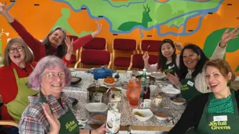 A group of adults sit around a table during a Joyfull Greens cookery session, with bowls of food, utensils and a brightly-coloured mural on the wall behind them. They are waving and holding their hands in the air, looking excited and happy.