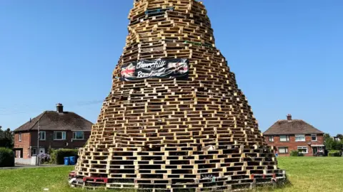Close shot of bonfire, built using wooden pallets, at Churchill Park. There are pallets on the grass in the foreground. In the background, there are two semi-detached, red brick properties.