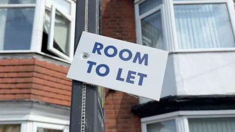 Room To Let sign in blue capital letters on a white background in front of terraced housing in Hull