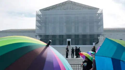 People hold rainbow umbrellas on the steps of the US Supreme Court building on a sunny day. 