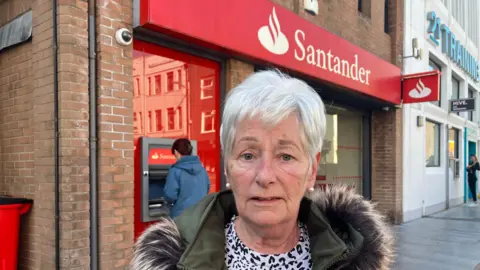 A whit haired woman stands outside the Santander Strabane branch. She has pearl earrings and a parka.