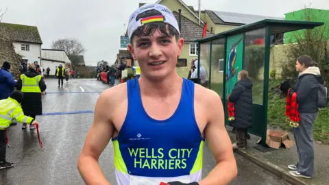A 17-year-old boy wearing a bright blue and neon green Wells City Harriers tank top and a white cap facing backwards. He has brown hair which is poking out from beneath the cap and his mouth his slightly open after just winning a race. He is stood on the middle of the road in a Somerset village on a grey and damp winters day. Spectators can be seen behind him, some near a bus stop at the side of the road, waiting to cheer other participants on.