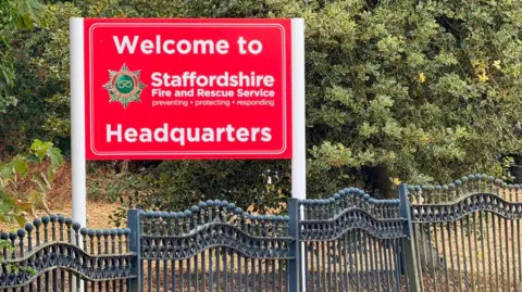 BBC A red sign with white lettering which says 'Welcome to Staffordshire Fire and Rescue Service headquarters'. There is a black railing beneath the sign and trees behind.