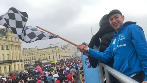 Olly Alexander waves a black and white checked flag at the start line of the Brighton Half Marathon 2026. He is wearing a blue hoodie which says 'The Sussex Beacon'.