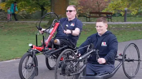 The men are both smiling as they travel on a path in a park on handcycles