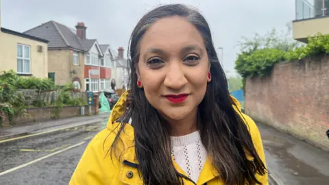A woman wearing a yellow rain coat stood in the street smiling into the camera