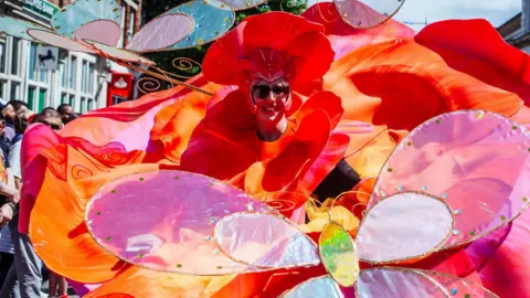 Aleksandra Warchol A woman, dressed like a giant flower, taking part in a carnival parade. She is wearing a large red hat, and has petals and a butterfly, made out of fabric, in front of her. She is smiling and has on sunglasses.