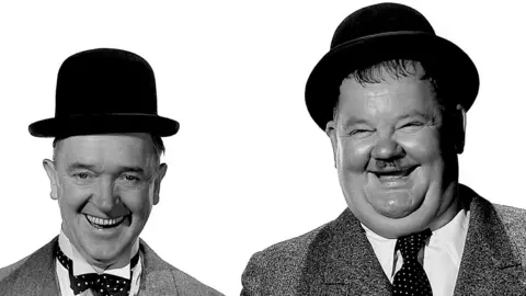 Getty Images A black and white photo of Laurel and Hardy wearing their iconic Derby hats and smiling broadly. Laurel, on the left, is slim and wears a bow tie. Hardy is larger and wears a straight tie.