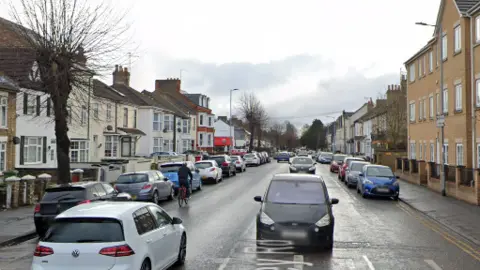 Google A street view image of the road with cars parked at either side and other cars traveling in both directions down the centre. There are houses at either side of the street. 