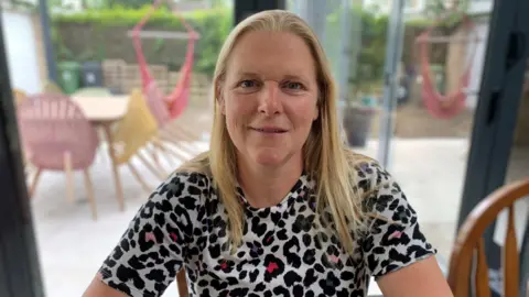 Une femme au début des années 50 était assise à la table de la cuisine portant un t-shirt à imprimé animal coloré avec des cheveux blonds à longueur d'épaule souriant.