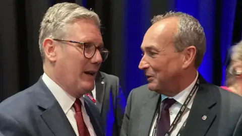 Sir Keir Starmer and chief minister Lyndon Farnham. Sir Kier is wearing a suit, glasses and a red tie.Farnham is wearing a dark grey suit and a grey tie. They are laughing together.