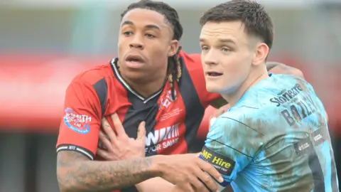 Isiah Noel-Williams in red and black Kettering shirt grapples with a Doncaster Rovers player in muddy sky blue shirt