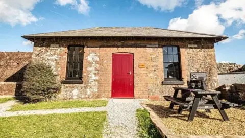 The Straw Store building which is a brick and stone building with a red front door and two windows on either side. There is a bush to the left of the building and a picnic table to the right.