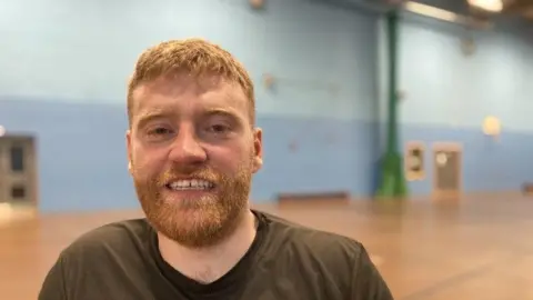 BBC A man with ginger hair and beard is in a sports hall, he is smiling at the camera and wearing a grey t shirt. 