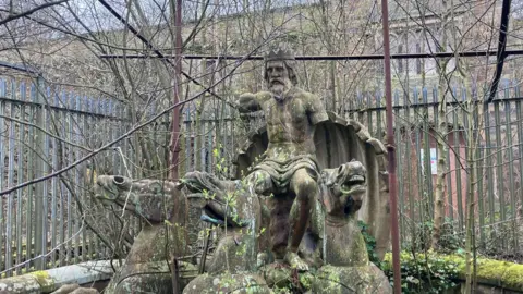 Photograph of the Neptune Fountain in the Stable Courtyard at Winstanley Hall. The overgrown, moss-covered and vandalised statue has its arms missing. 