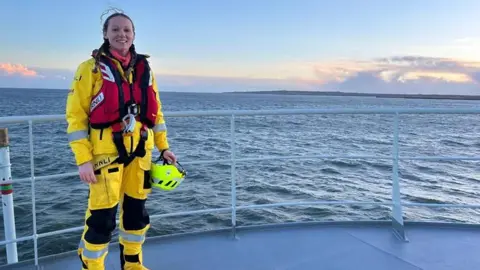 Sheridan Bowler standing on the deck of a ship. She is wearing yellow wet-weather clothing and a red lifejacket while holding a yellow helmet. Her dark hair is tied in plaits with the wind flicking strands on top of her head. The sea has lots of small waves on the surface and the coastline can be seen in the distance.