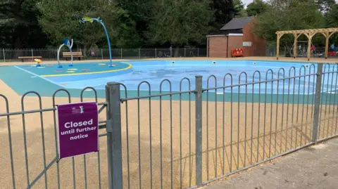 A fenced-off children's paddling pool in a park, with splash pad including mini jets. It is surrounded by green and yellow flooring, with benches around the outside, and trees can be seen beyond. A purple sign on the gate says closed until further notice.