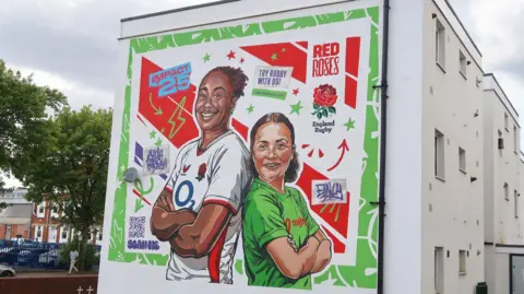 A mural on the side of a building. The wall is white and the painting features a woman in a white England rugby top and a girl in a green rugby top.