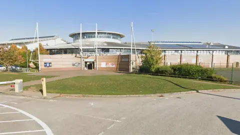 Google Street View image of the outside of Grantham Meres Leisure Centre, with light brown walls and a large, round lantern roof in the middle of the building