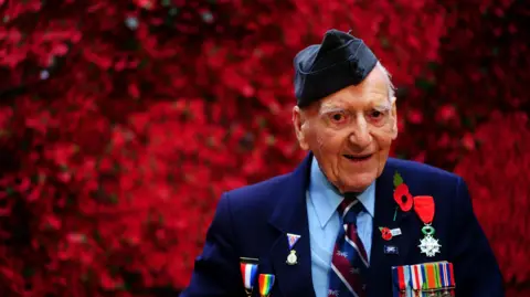 PA Media Bernard Morgan, an older man, wearing a suit has numerous medals on his chest and Poppy Appeal pin badges. He is positioned against a vivid red backdrop.
