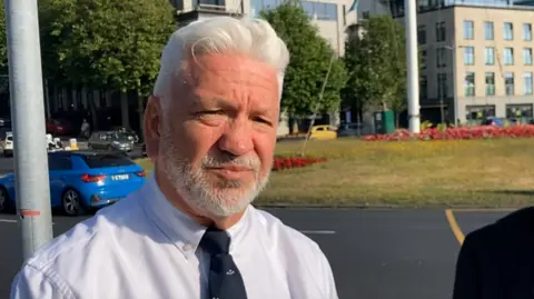 Calum is pictured in St Peter Port. A roundabout is visible behind him. He has slick white hair and is wearing a shirt and tie. He has a grey beard and is smiling. 