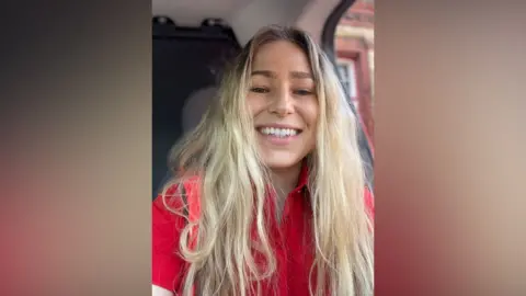Sitting in a vehicle and wearing a red top, Katy Davies smiles at the camera. She has long blonde hair.