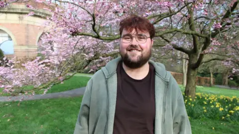 The image shows a young man standing outdoors in a park during springtime. He is smiling at the camera and wearing glasses, a dark t‑shirt, and a green hooded jacket. Behind him, pink blossom-covered trees are in full bloom, with green grass and yellow flowers visible at ground level.