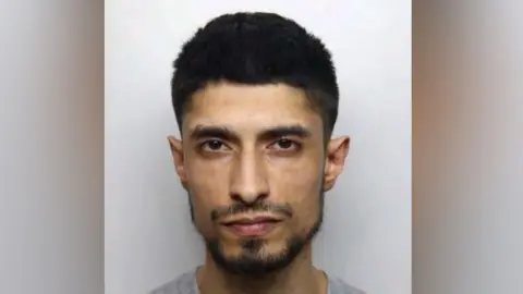 Yasar Hussain: A dark-haired man with a dark beard wearing a grey round-necked t-shirt. He is appearing in his police custody photo.