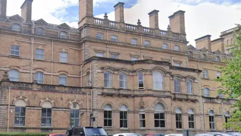 Google A wide hotel front with distinctive historic architecture. The top of some parked cars be seen in the foreground.