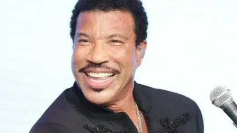 Lionel Richie is in a black bedazzled suit standing beside a microphone. 
He has short, curly, black hair and facial hair. He is wearing a large silver chain. 