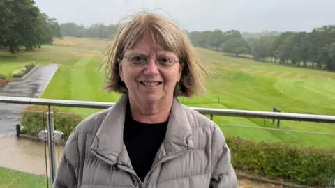A middle-aged woman in glasses looks at the camera. Behind her is a golf course