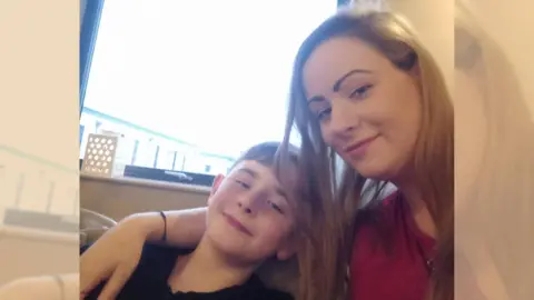 Hannah Foster A woman and boy sitting on a sofa. The woman has her arm on the shoulder of the boy and they are both smiling.