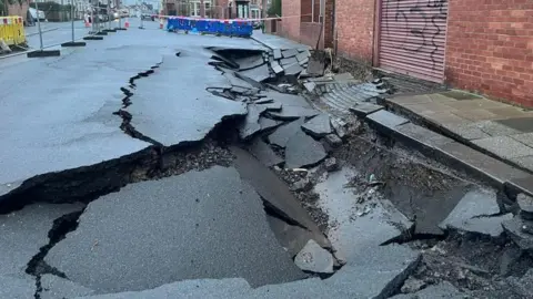 Grey temporary fencing and fluorescent barriers surround the sinkhole, pictured in January. The grey tarmac road is splintered and sunken.
