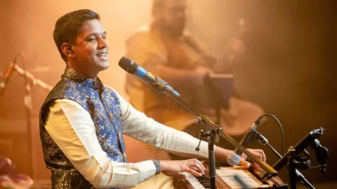 Wolverhampton Grand A man singing into a microphone. He is playing a keyboard and is wearing a dark blue and gold top with white sleeves showing underneath. 