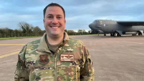 BBC Lieutenant Colonel Jared Patterson wearing his US Air Force camouflage uniform, smiling at the camera as a B-52 bomber stands on the runway behind him