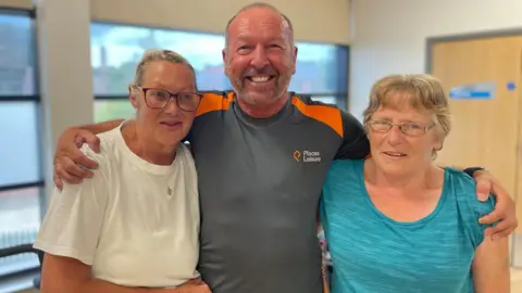 Simon Thake A man in a grey sports top hugs two nervous looking women both in sports tops and wearing glasses