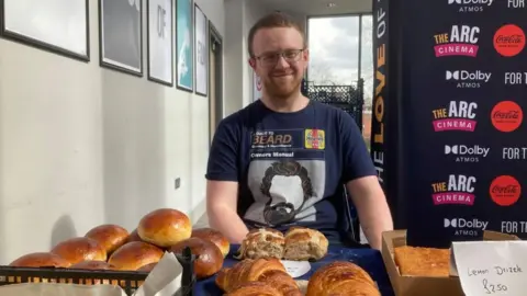 Martin Heath/BBC Ben Welch with short ginger hair and beard behind a selection of baked products on his stall