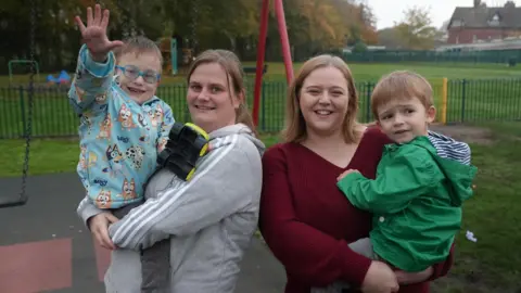 BBC Linzi and her son Roni and Courtney and her son Charlie. The boys are in their mums' arms and they're all standing in a play park in front of some swings. Roni is waving at the camera. 