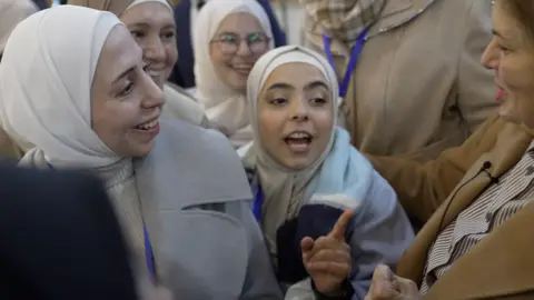 A group of young female students in light-coloured headscarves smiling as they interact with Hind Kabawat. One of the students is speaking and pointing her finger for emphasis. Kabawat is wearing a brown coat and striped shirt, and is smiling. 