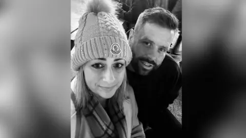 Charlotte Busby Black and white photo of a man and woman, Charlotte and Matthew. Charlotte's sparkly bobble hat reads 'Moncler' and she is wearing a coat and scarf. Matthew sits behind her, he is wearing a dark jumper, has a short beard and a quif hairstyle.