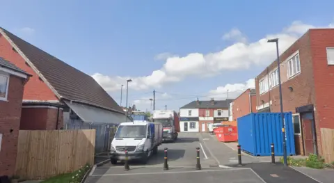 A road with bollards in the foreground and buildings wither side. There are two lorries parked on the left